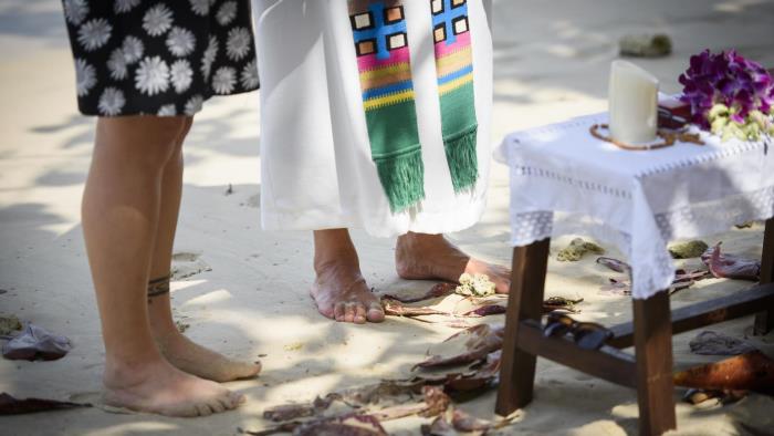 Två personer står bredvid varandra vid ett lågt altare på en sandstrand. En av dem har prästkläder. 