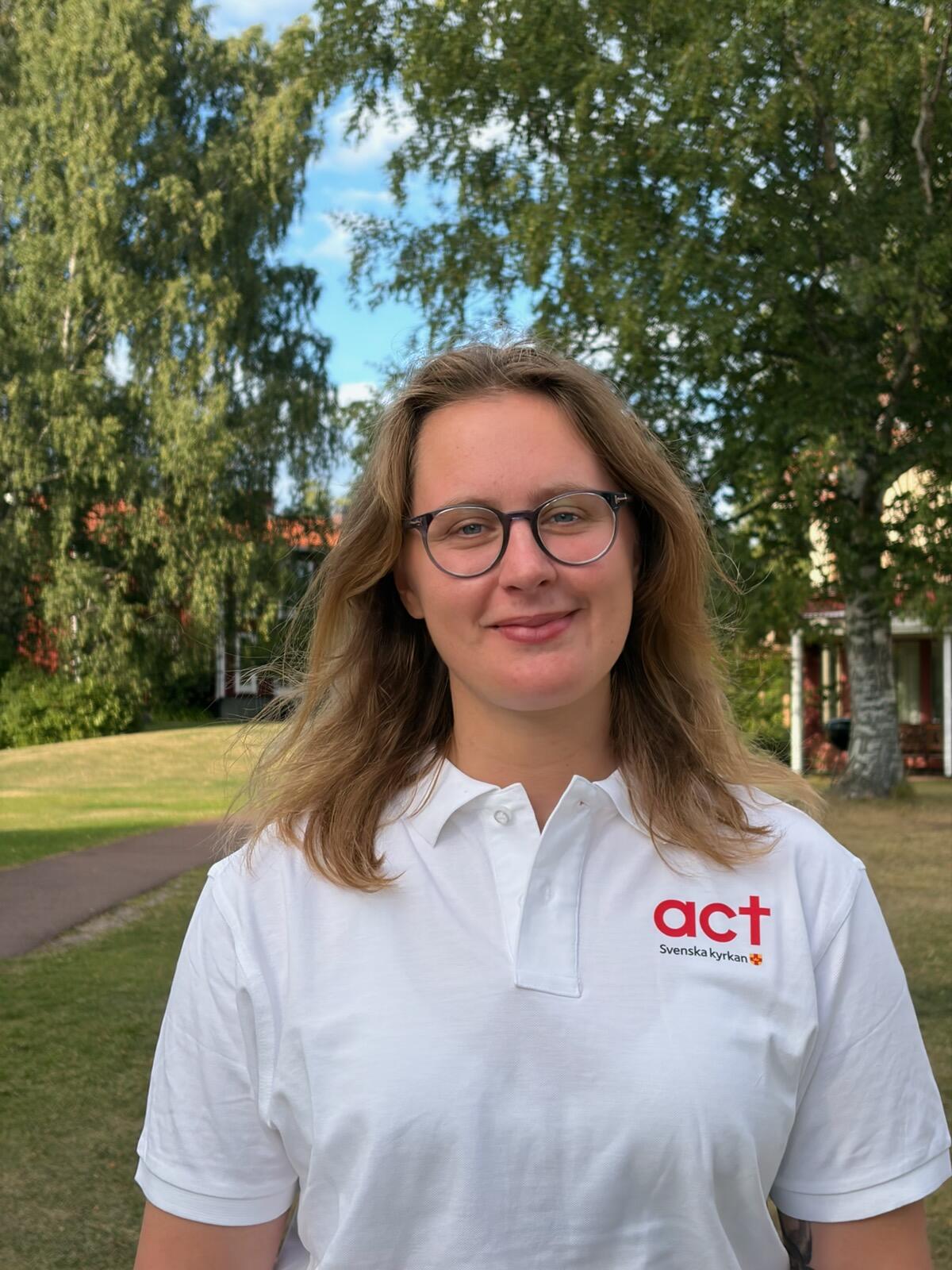 Porträttbild på Amelia von Schantz. Hon står utomhus och bär en vit pikétröja med Act Svenska kyrkan-logga.
