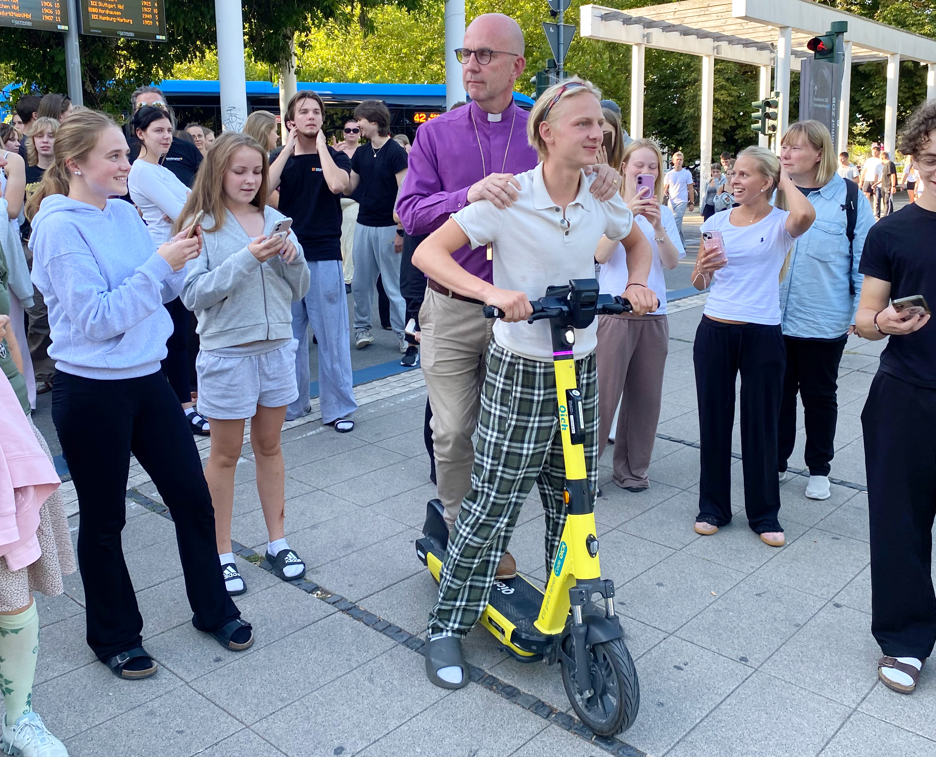 En ung kille står längst fram på en elsparkcykel, biskopen står bakom honom och håller honom på axlarna. De är omgivna av ungdomar som ler eller tittar i sina mobiltelefoner.