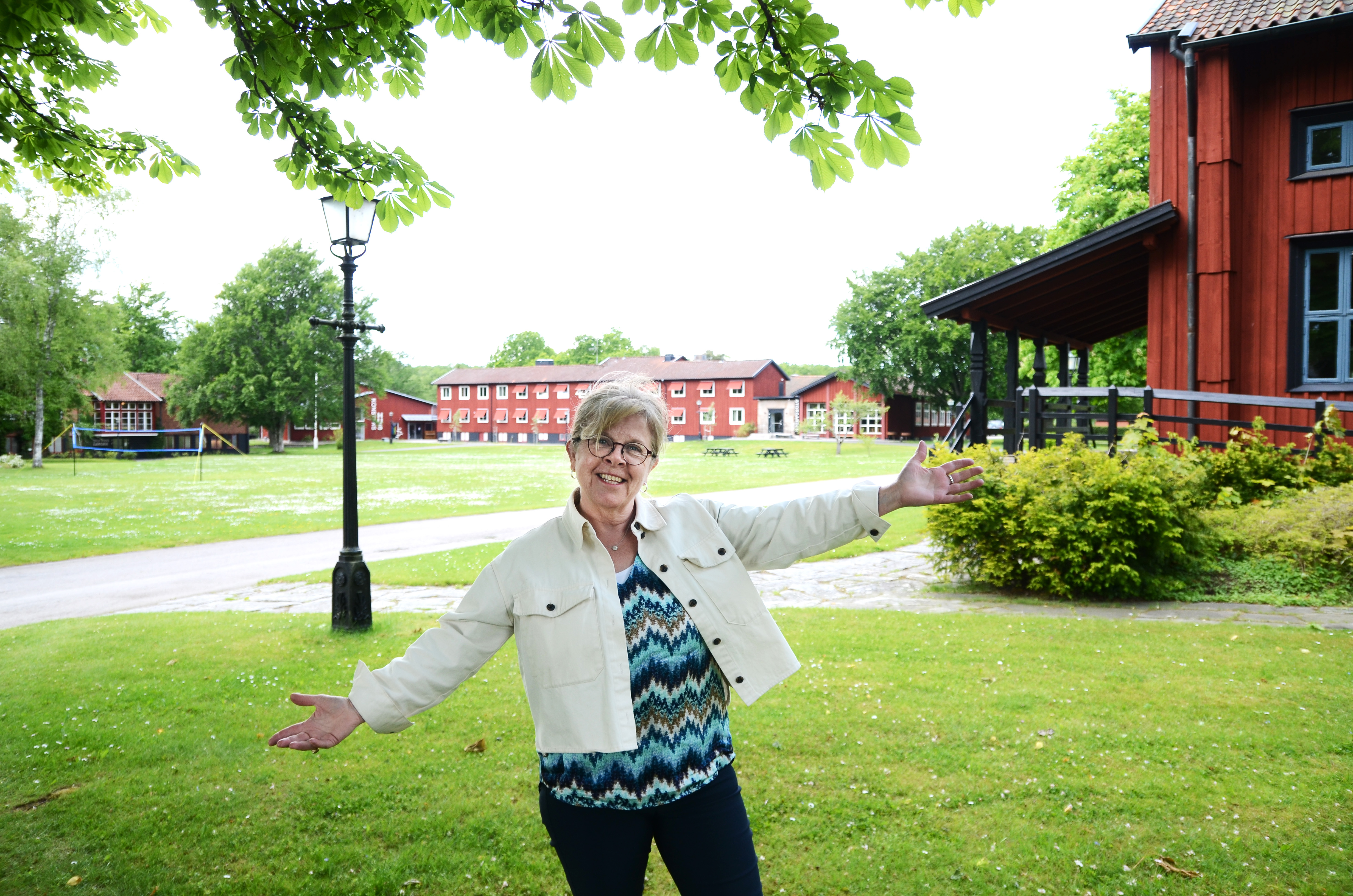 Marita Nilsson håller välkomnande ut armarna. I bakgrunden syns flera av skolbyggnaderna, den stora gräsplanen och en del av kapellet.