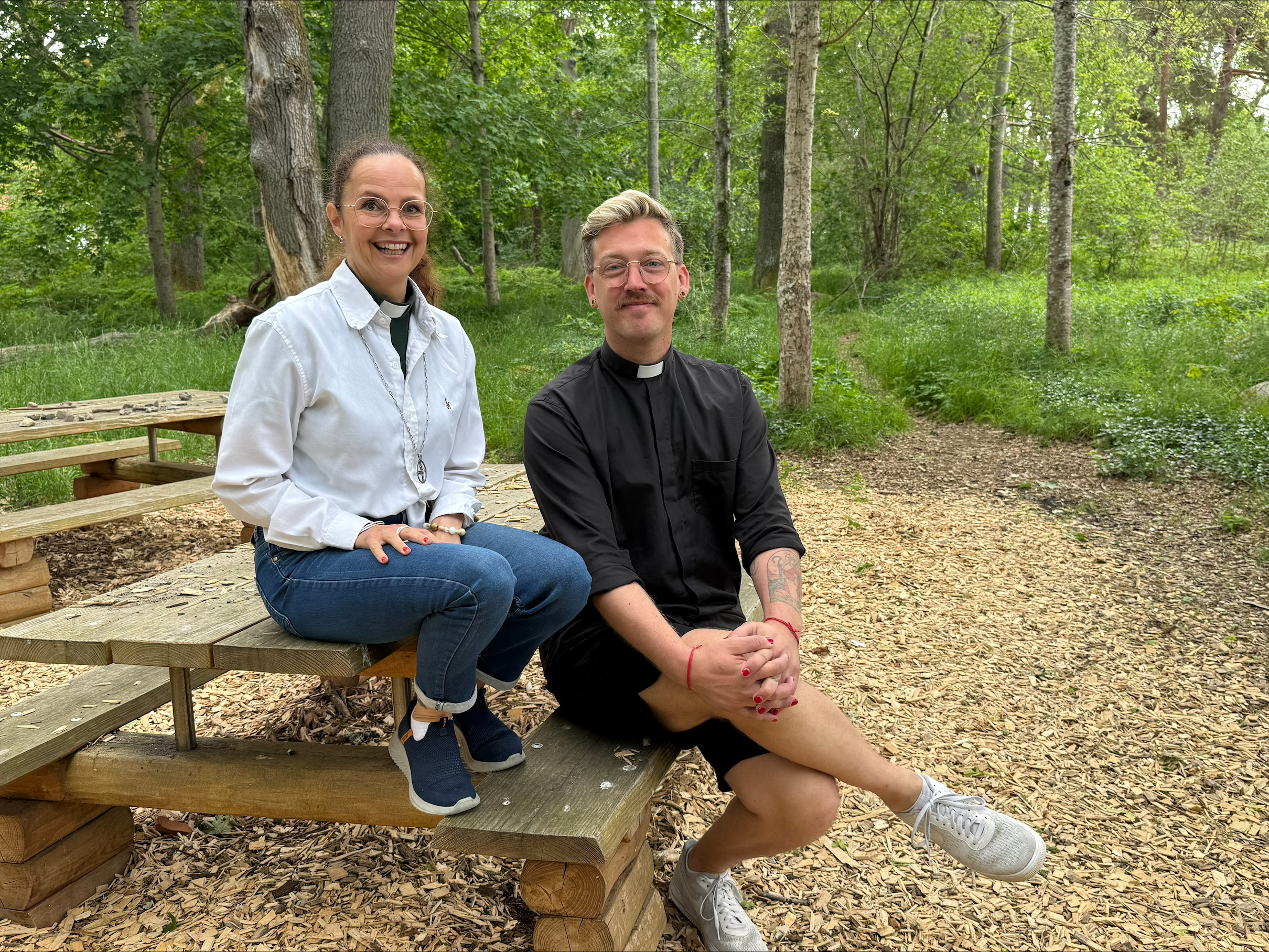 Diakonen Malin Hjertquist sitter på ett picknickbord, prästen Erik Horner sitter på bänken intill. De är omgivna av gröna lövträd.