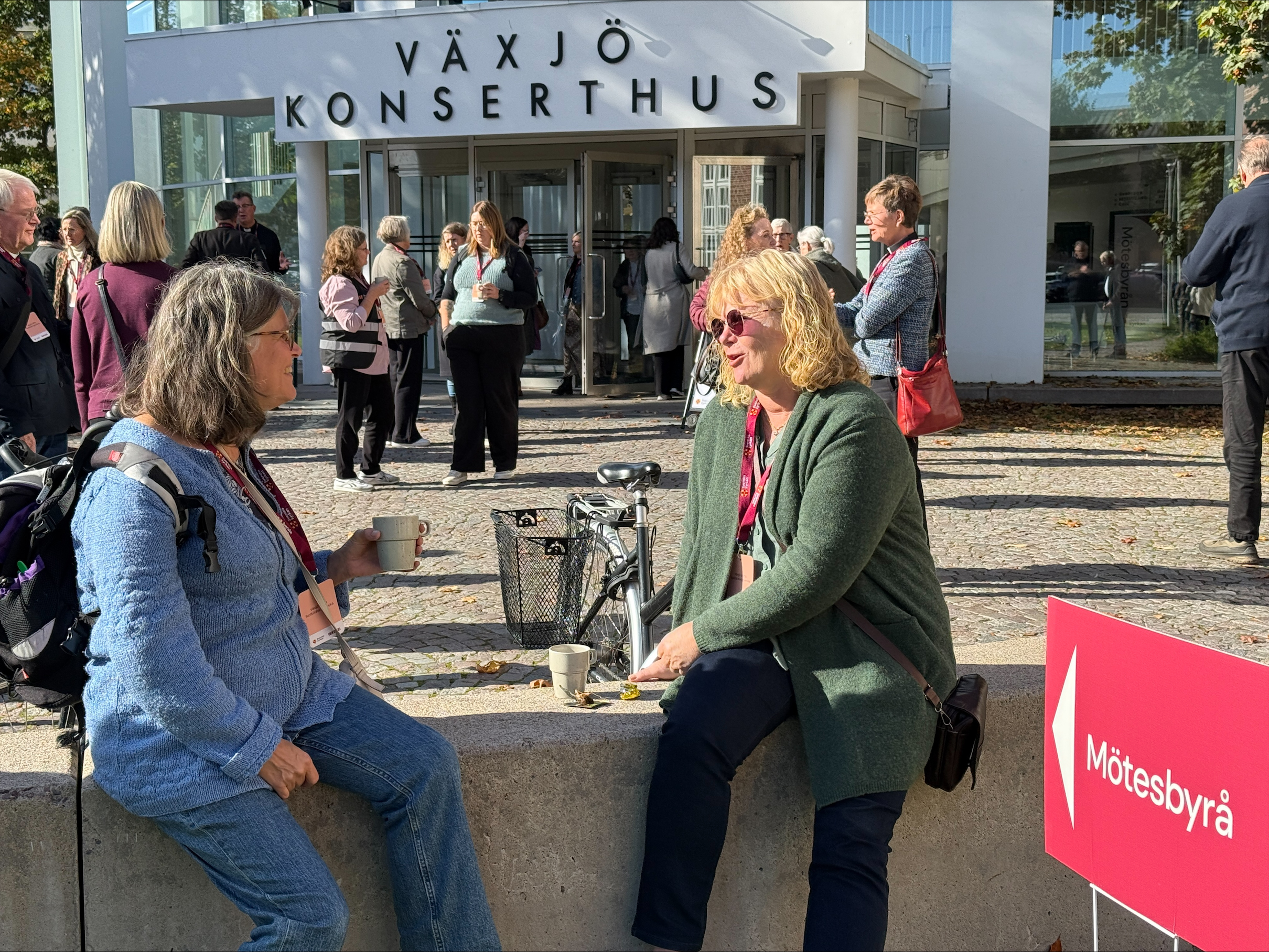 Två kvinnor sitter på en låg mur och samtalar. I bakgrunden syns Växjö konserthus och fler människor som samtalar.