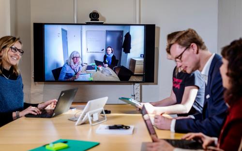 Foto på personer som sitter med laptops runt ett konferensbord. På väggen en bildskärm där ytterligare personer sitter runt ett konferensbord. En del ser glada ut, andra koncentrerade.