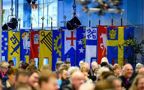 Bild från Kyrkomötet med ledamöter i förgrunden och flaggor i bakgrunden.
