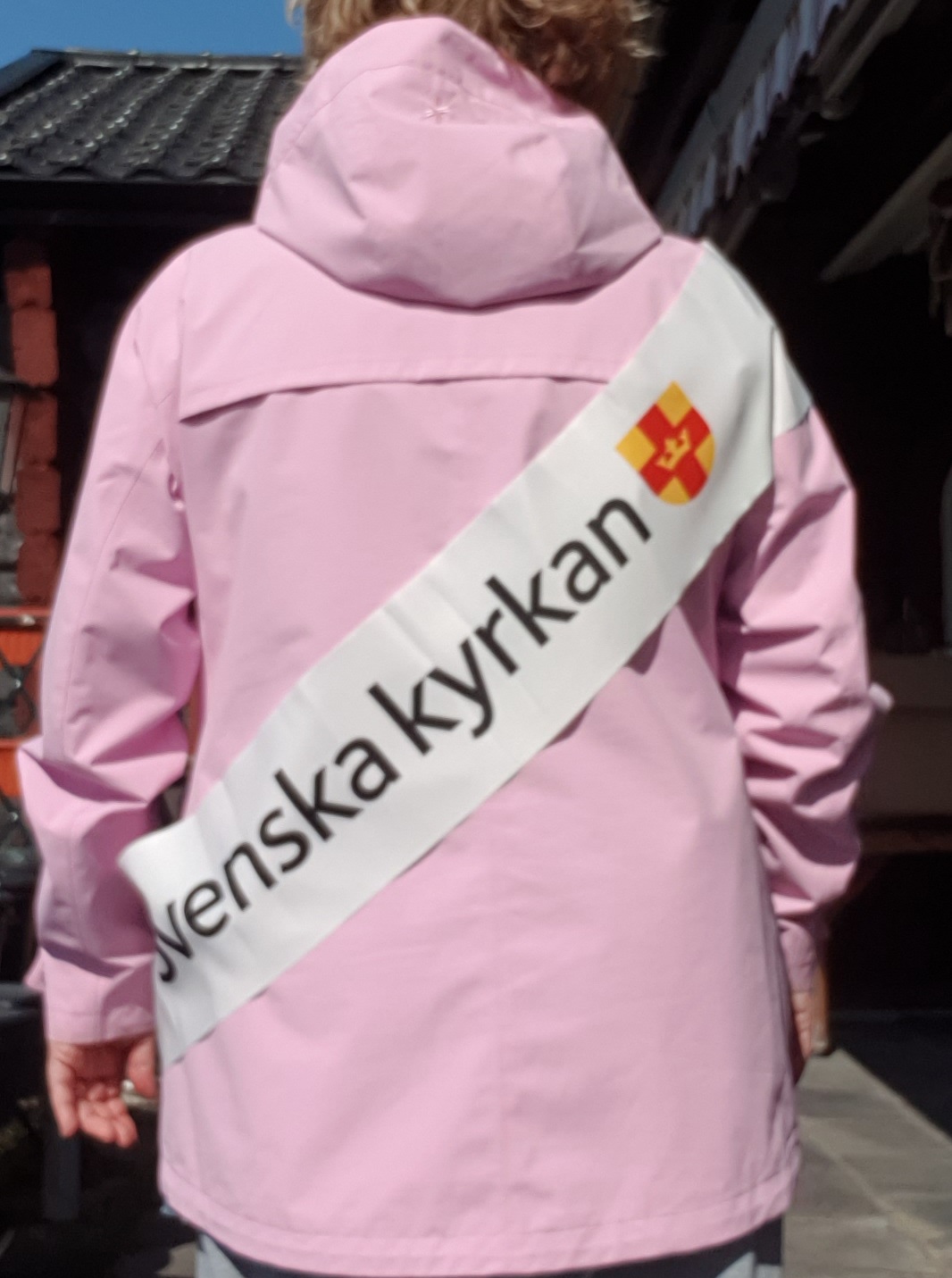 Foto på en person i rosa jacka fotograferad bakifrån. Personen bär en banderoll med Svenska kyrkans logga över axeln.