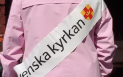 Foto på en person i rosa jacka fotograferad bakifrån. Personen bär en banderoll med Svenska kyrkans logga över axeln.