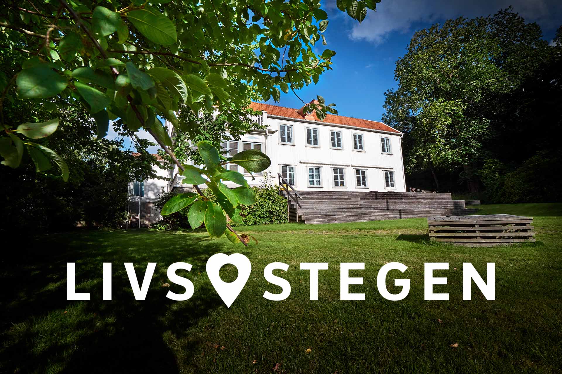 En vit stor prästgård under blå sommarhimmel. Gröna buskar i förgrunden.