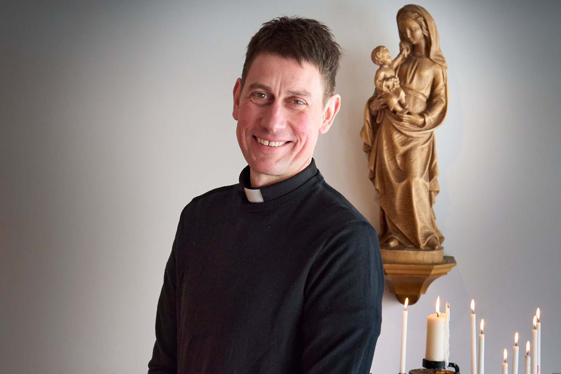 Bilden föreställer Henrik Lange i prästskjorta, ståendes bredvid en ljubärare med tända ljus och framför en mariastaty av trä som hänger på väggen bakom.