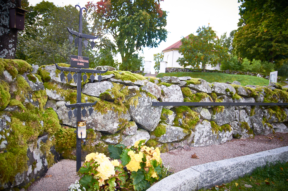 Ett kors med tre korsarmar i smidesjärn. Står framför en stenmur med mossa. I bakgrunden Härryda kyrka.