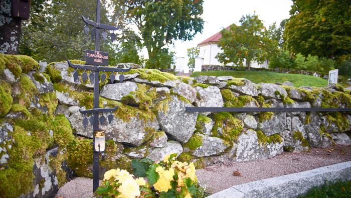 Ett kors med tre korsarmar i smidesjärn. Står framför en stenmur med mossa. I bakgrunden Härryda kyrka.