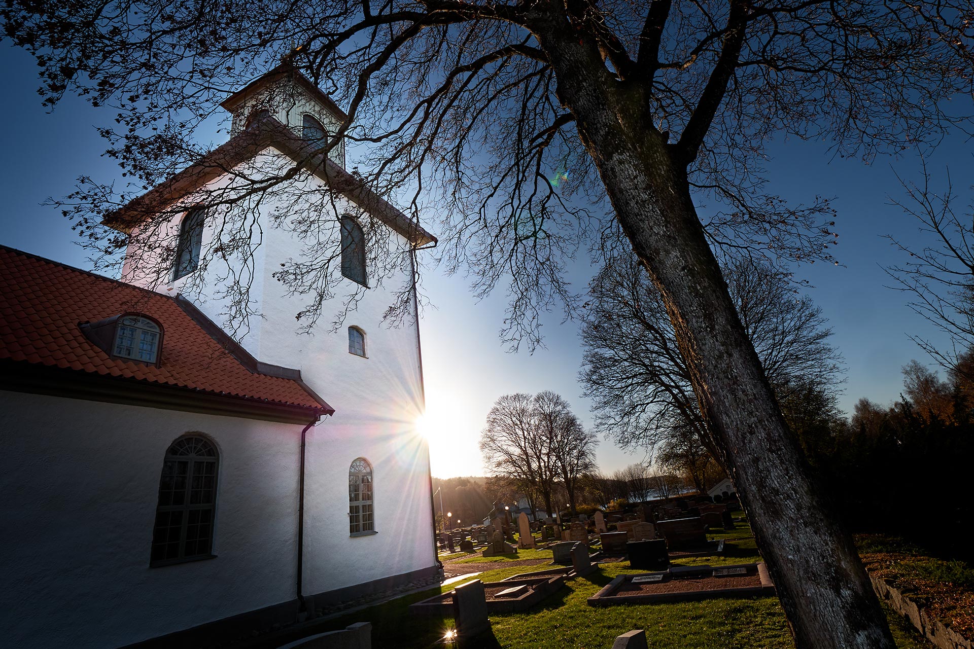 En vitkalkad 1830-talskyrka. Fotad i motljus. Ett träd till höger i bild.