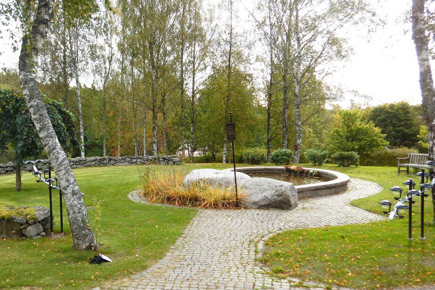 Minneslunden på Härryda kyrkogård. En stenomgärdad granitfontän med två stora stenblock framför. I förgrunden en björk.