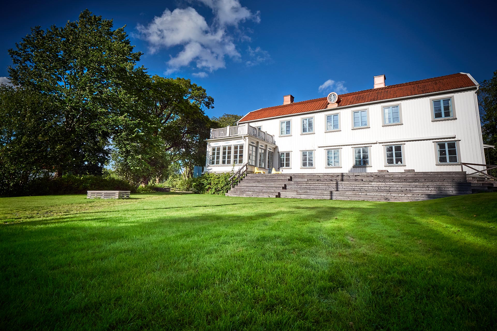 En vit herrgårdlik träbyggnad. Framför huset en grön gräsmattar och stora träd.