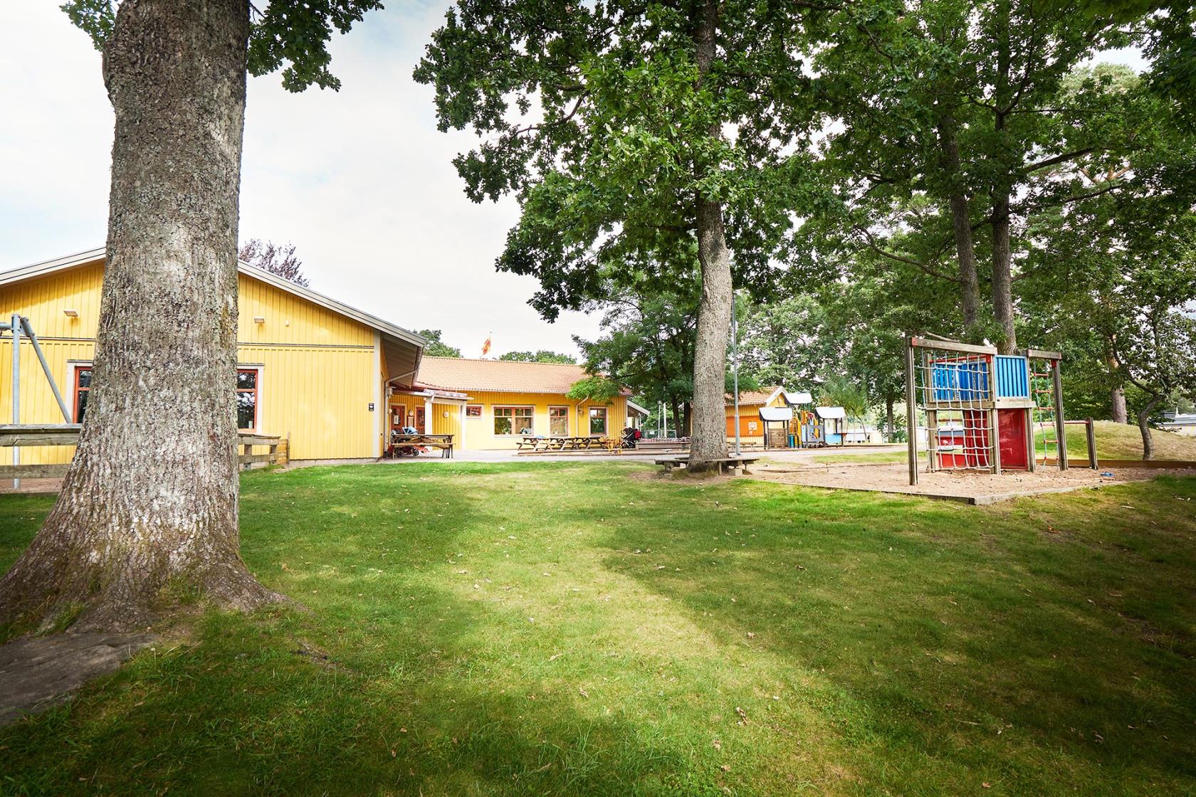 En gul träbyggnad, Solbackens förskola, omgiven av träd. På en gårdsplan sandlåda, klätterställning och lekredskap. 