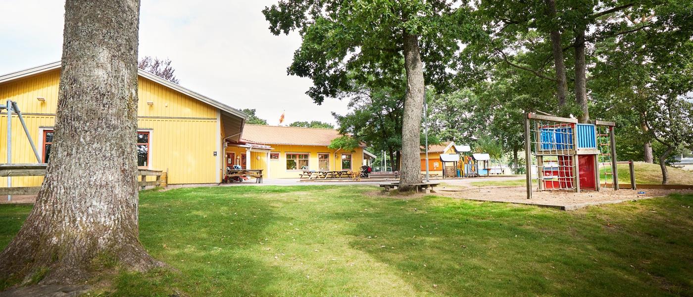 En gul träbyggnad, Solbackens förskola, omgiven av träd. På en gårdsplan sandlåda, klätterställning och lekredskap. 