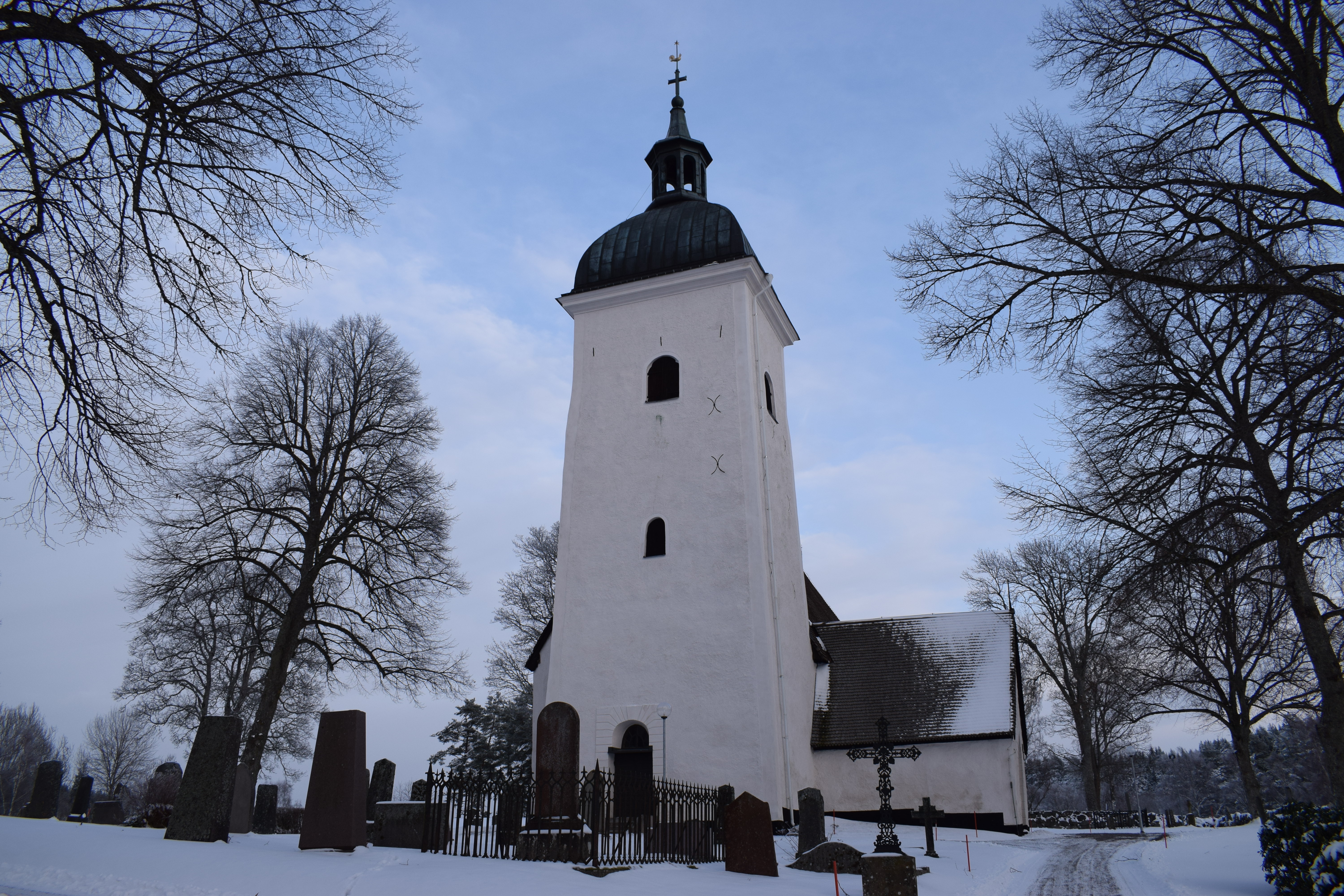 Vit, stenkyrka. Det höga tornet står i förgrunden. Snö på marken runtom. Gravstenar framför kyrkan.