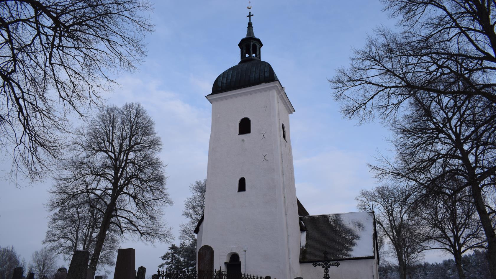 Vit, stenkyrka. Det höga tornet står i förgrunden. Snö på marken runtom. Gravstenar framför kyrkan.