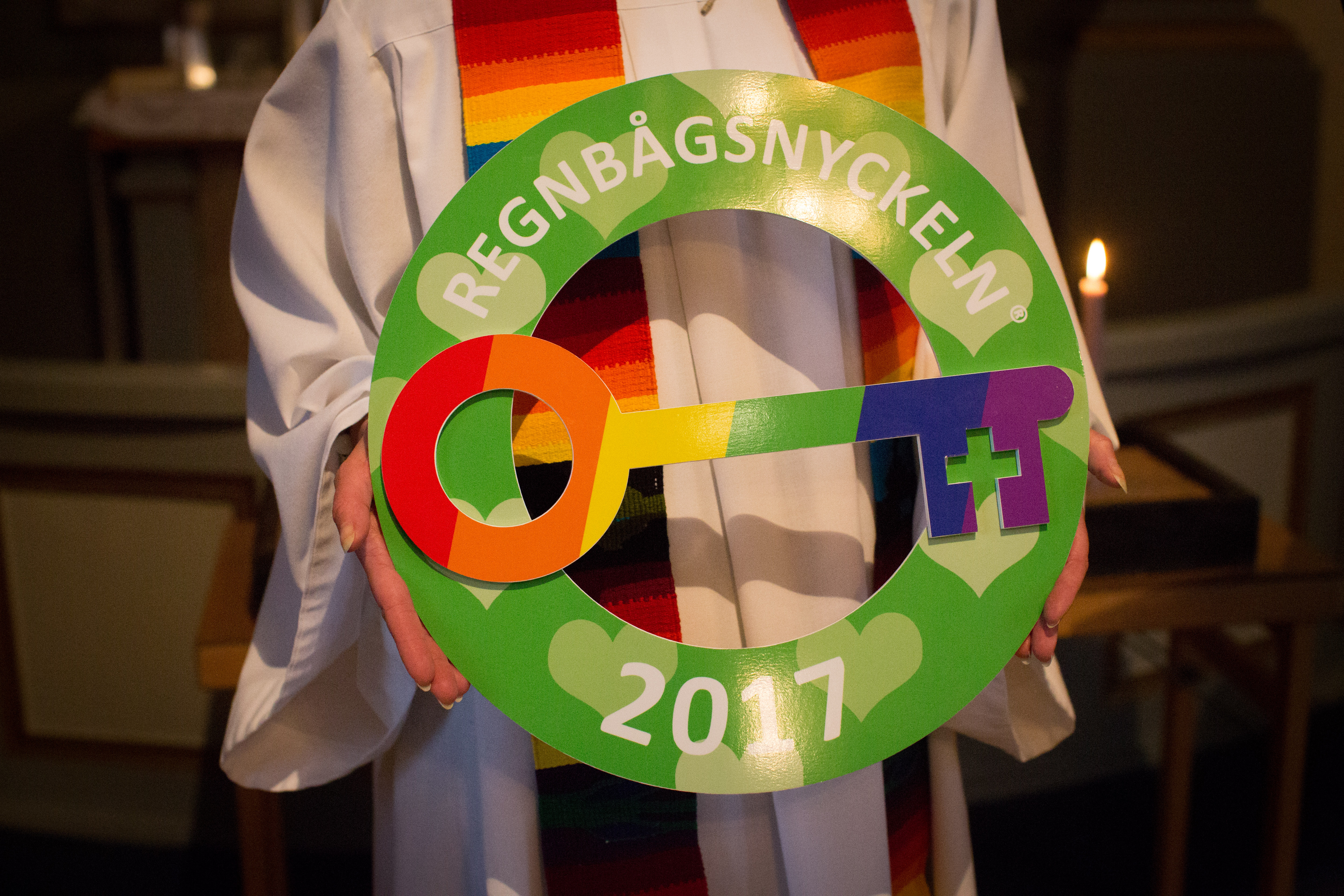 Präst håller upp den stora, färgglada regnbågsnyckeln från år 2017. Prästens stola är regnbågsfärgad. Prästen