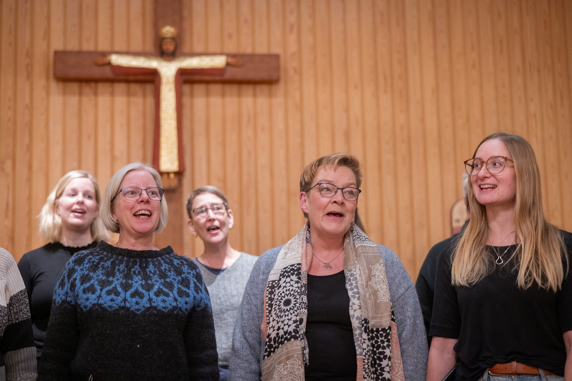 Kvinnor sjunger i kör. På väggen bakom den hänger ett kors. 