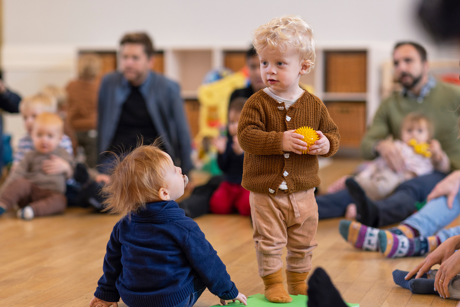 Barn med boll i handen. Runtomkring sitter vuxna och andra barn i en ring.