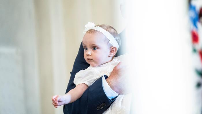 En bebis i dopklänning och hårband hålls i en mans armar.