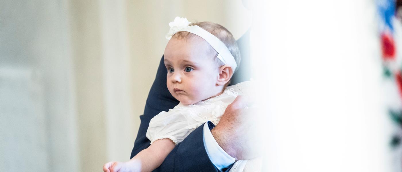En bebis i dopklänning och hårband hålls i en mans armar.