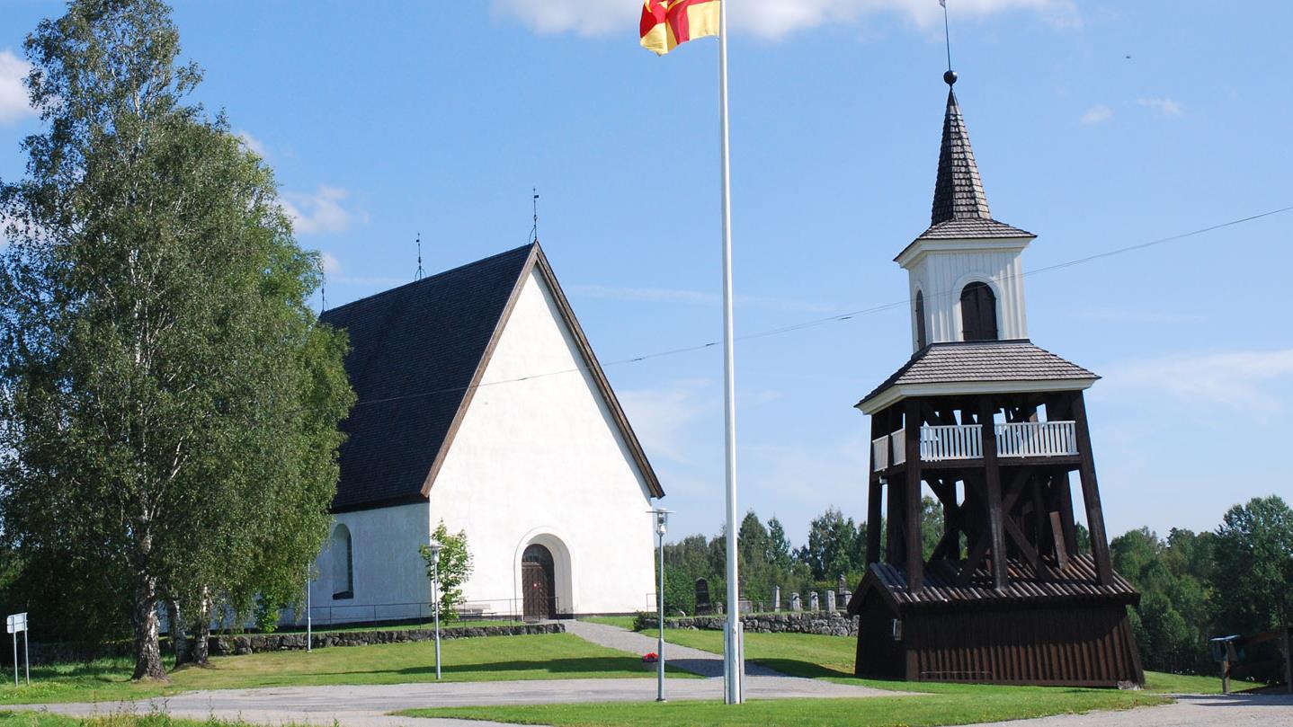 Överlännäs kyrka och klocktorn. Sommarbild. I förgrunden en flaggstång med Svenska kyrkans flagga hissad.