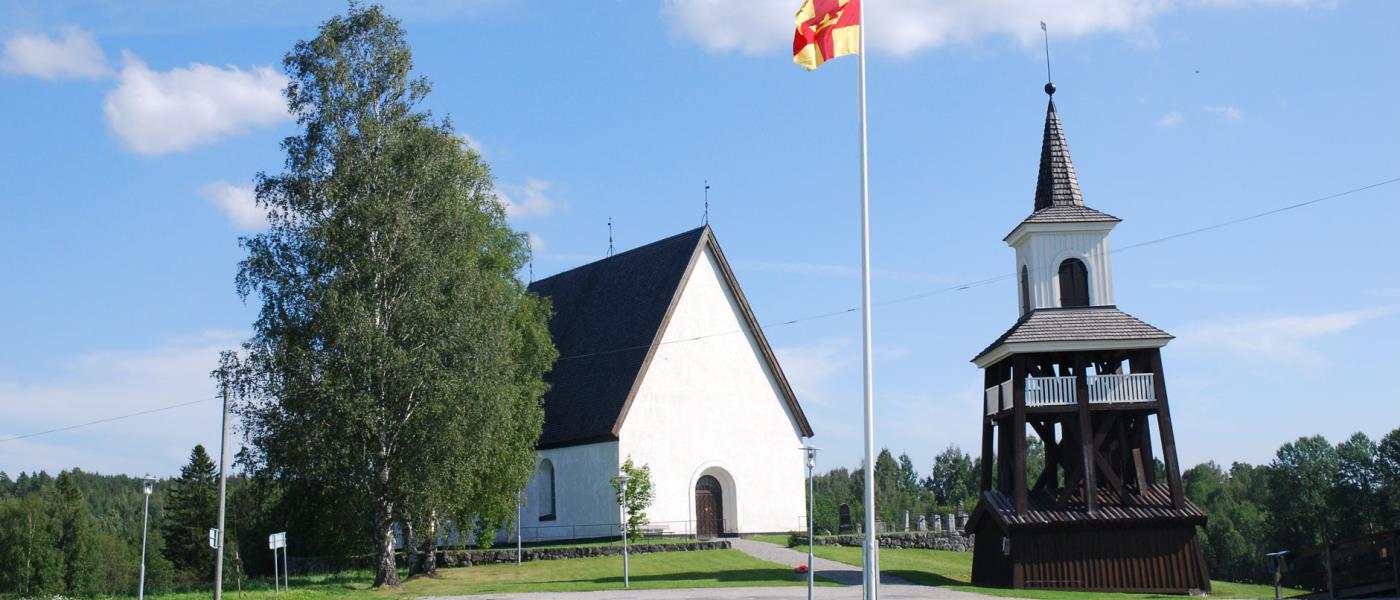 Överlännäs kyrka och klocktorn. Sommarbild. I förgrunden en flaggstång med Svenska kyrkans flagga hissad.