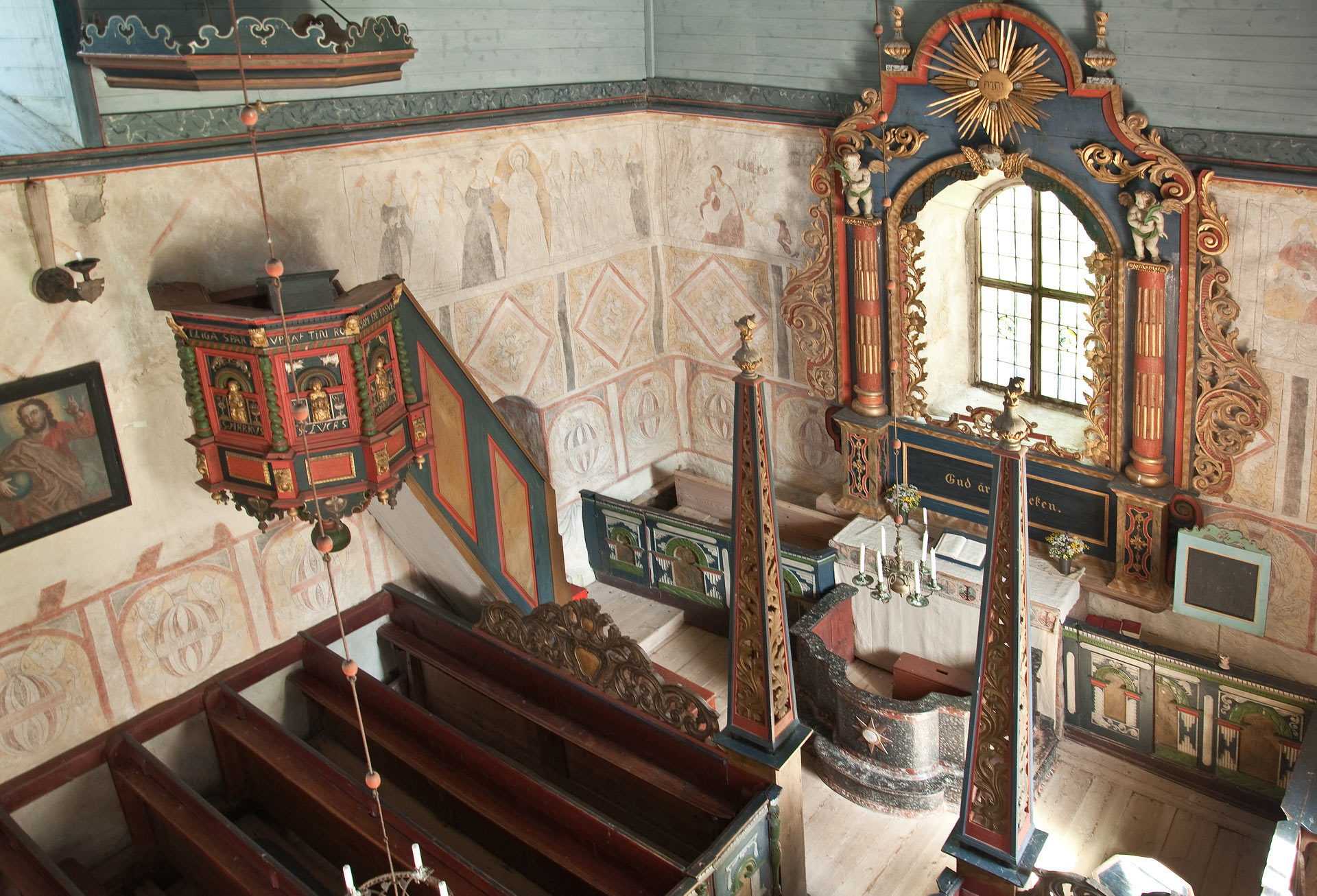 Interiör från Ramsele medeltida kyrka. Kor, altare, predikstol. Inredningen i trä går i rött,, blått, grönt och guld. På väggarna anar vi målningar.