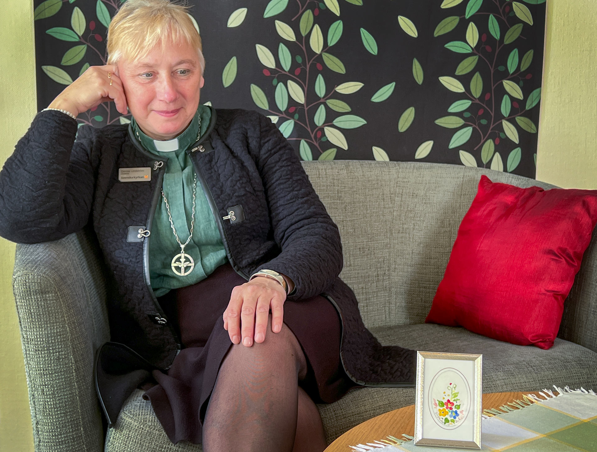 En kvinna med diakonskjorta sitter i en soffa. Hon tittar på en liten tavla med blomstermotiv.