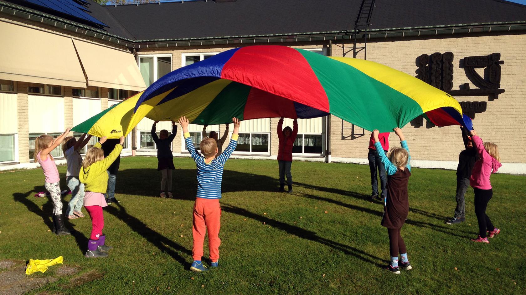 Tio barn och en ledare leker med en stor färggrann "fallskärm". De hjälps åt att kasta upp den i luften.De är utomhus på en gräsmatta.