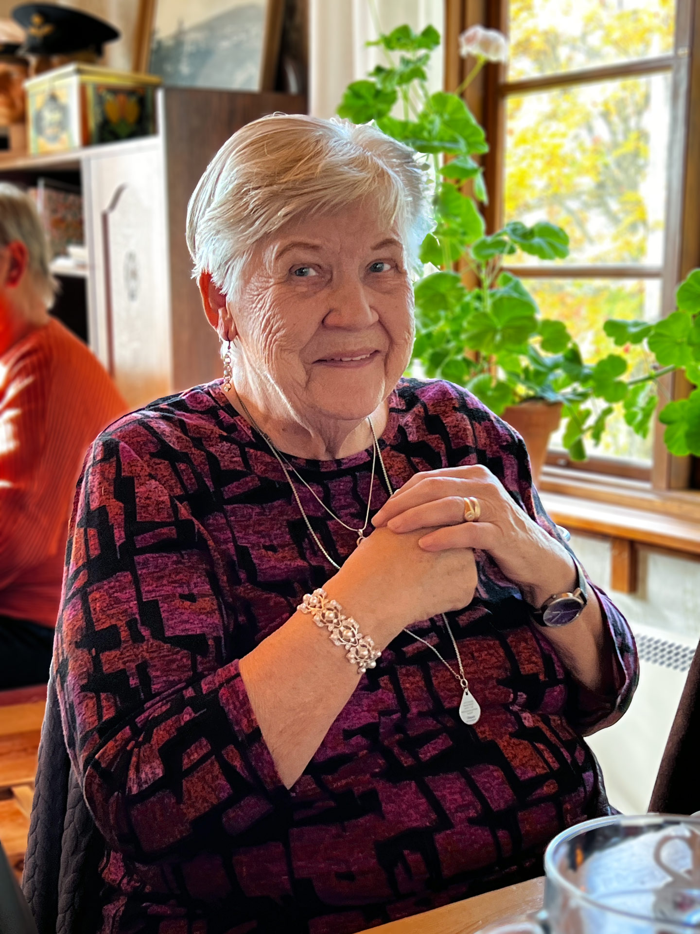 En äldre kvinna tittar in i kameran. Bakom är ett fönster ut mot höstfärgade björkar. Det står en pelargon i fönstret. I bakgrunden ser vi även att det sitter en kvinna till.