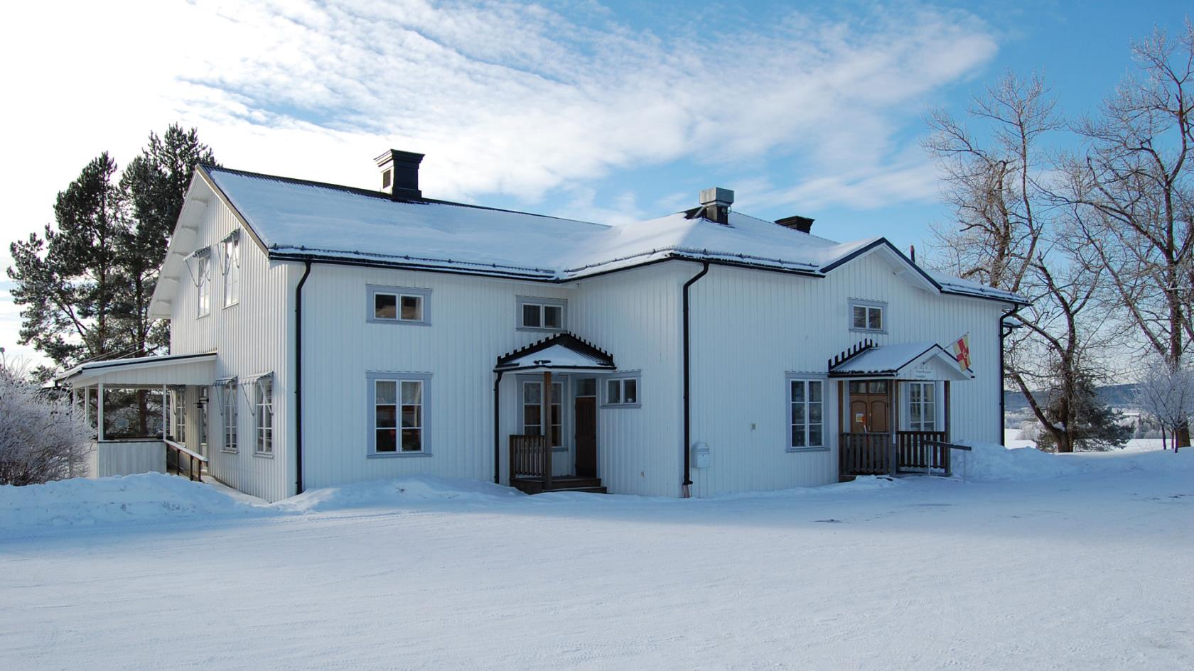 Långsele församlingshem. Vit träbyggnad. En vinterbild. Ovanför entrén hänger Svenska kyrkans flagga.