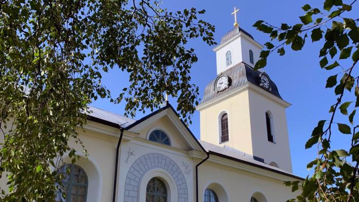 En stor stenkyrka med gulaktig puts. Det är sommar och vi ser kyrkan mellan gröna björkblad.