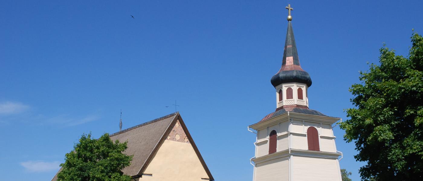 Boteå kyrka och klocktorn. I förgrunden delar av kyrkogården. Det är sommar. Mot den blå himmeln skymtar en svala.