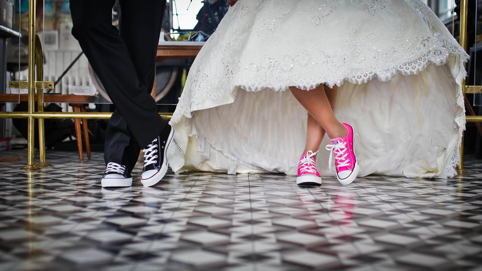 Ett brudpar fotograferat från knäna och nedåt. Vi ser klänning och kostymbyxor. På fötterna har de sneakers.