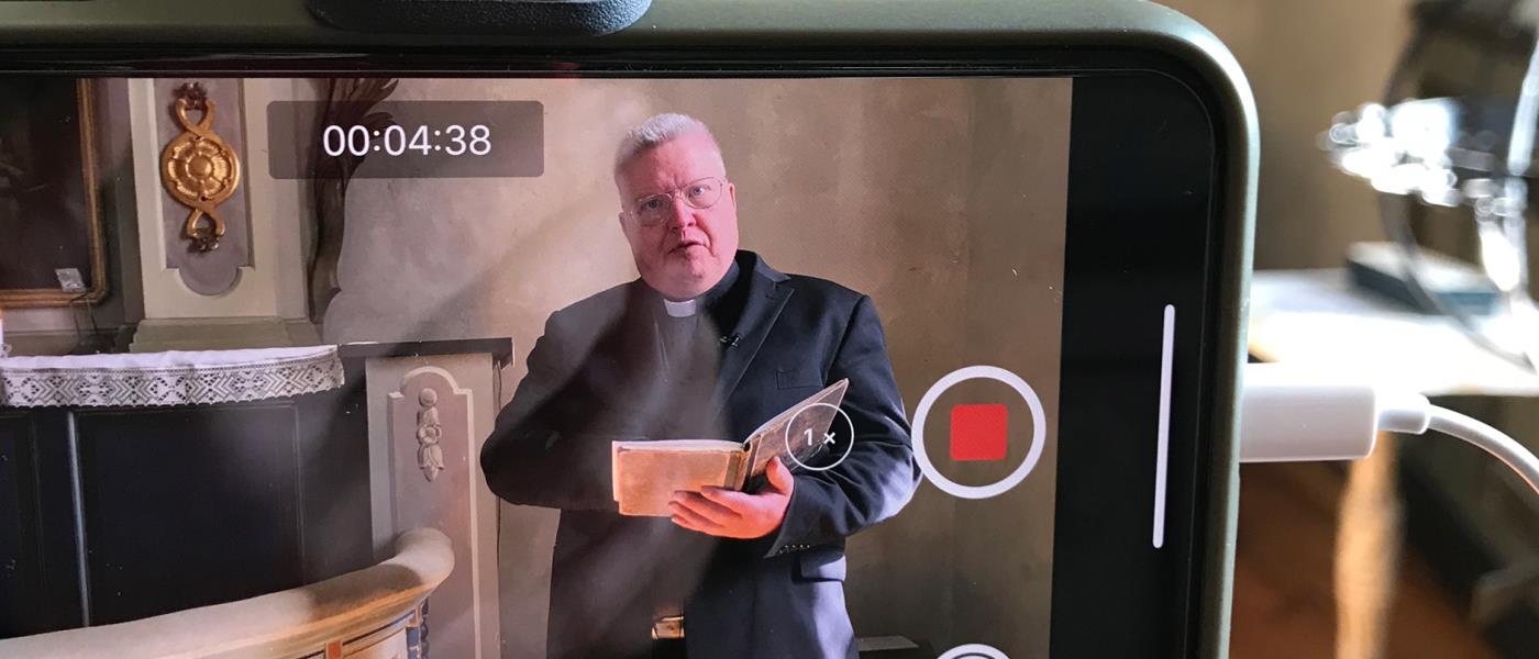 Genom skärmen på en mobiltelefon ser vi en präst som håller en andakt. 