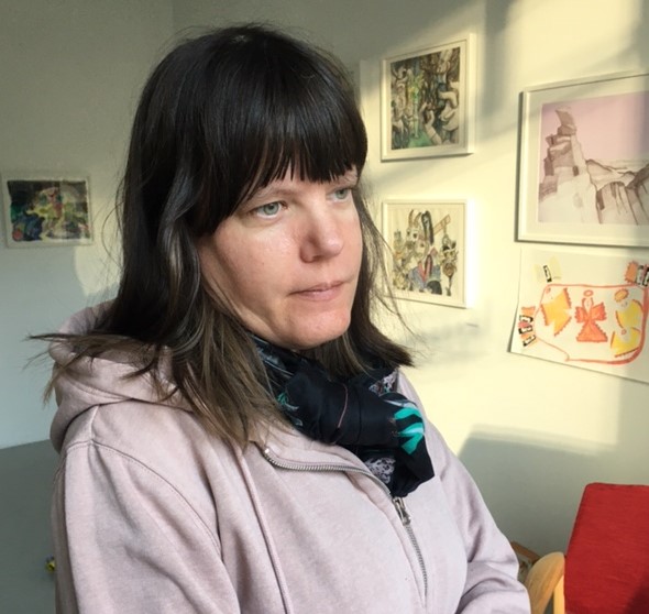 En porträttbild av konstnären Emma Asplund. I fonden hänger några av hennes konstverk.
