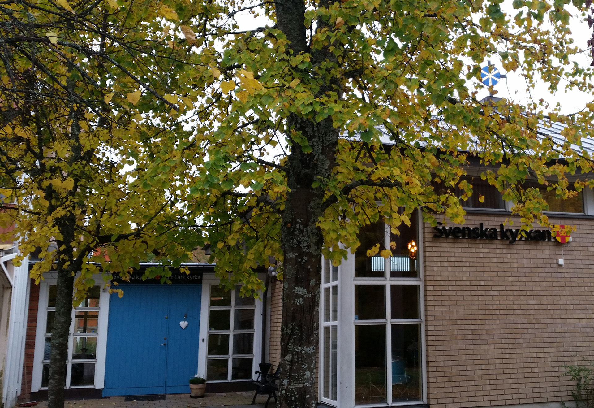 En gul tegelbyggnad med blå dörr. På huset sitter Svenska kyrkans logotyp. I förgrunden står två lövträd.