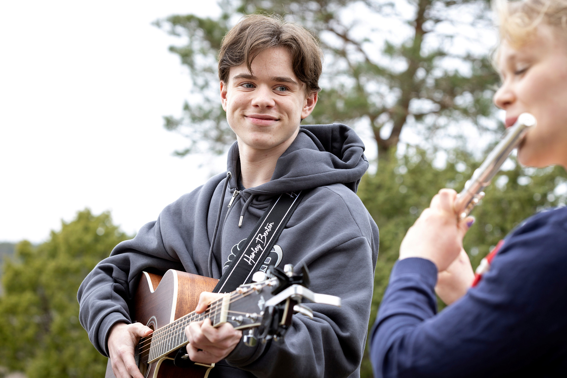 En ung kille står med en gitarr om halsen och ler varmt mot en tjej med blockflöjt i förgrunden. 