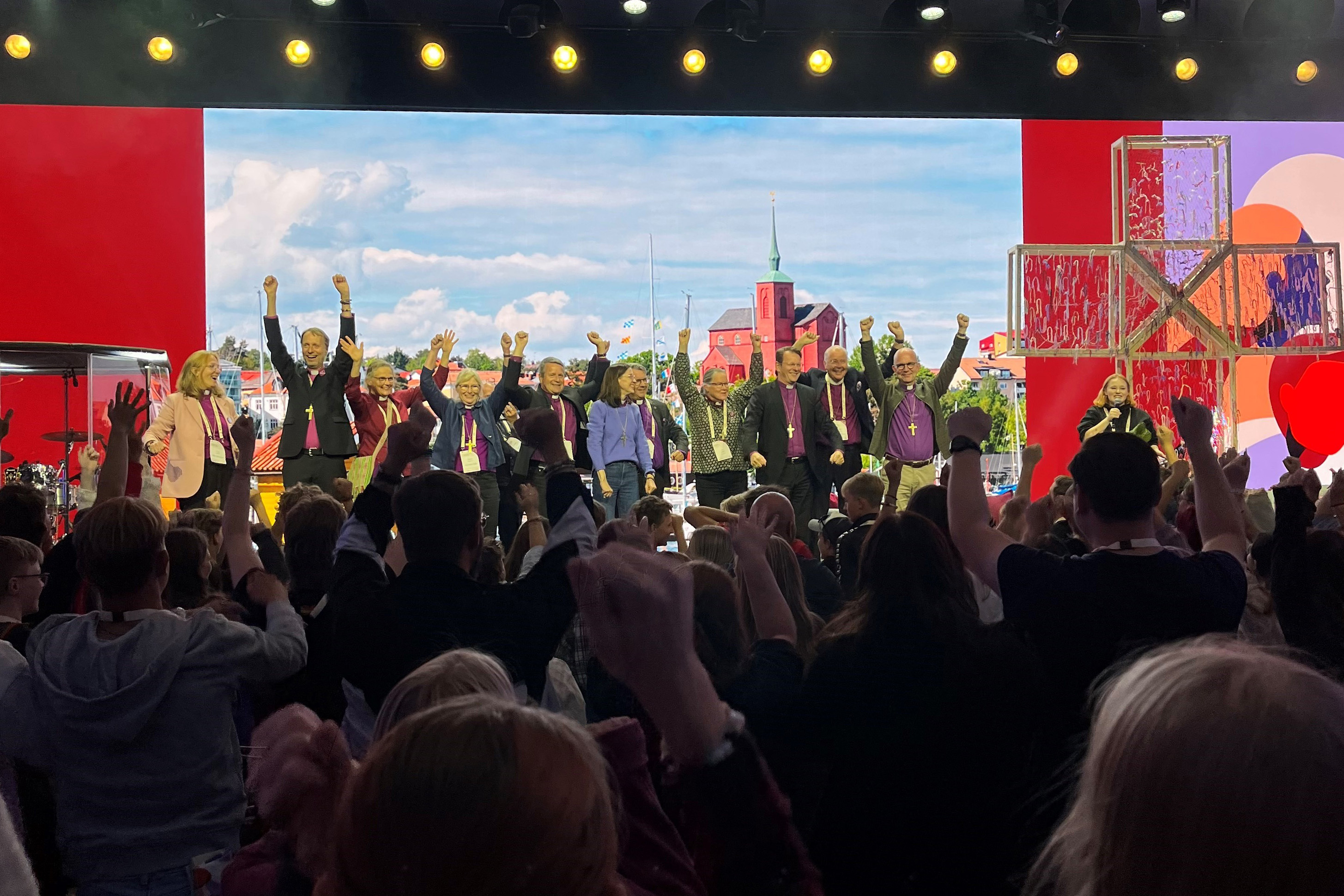 På en scen står biskoparna och viftar med händerna. Framför den finns ett publikhav med ungdomar som gör samma sak.