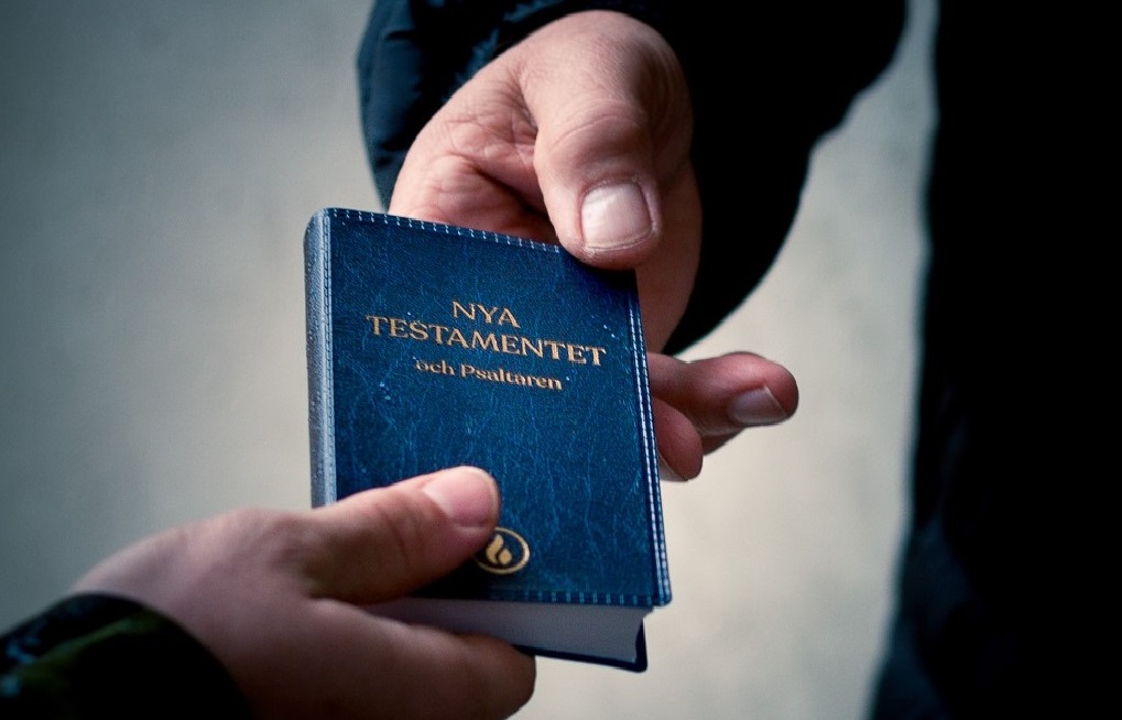 Närbild på två personers händer där den ena räcker över en liten blå bok med texten ”Nya testamentet och Psaltaren” till den andra.