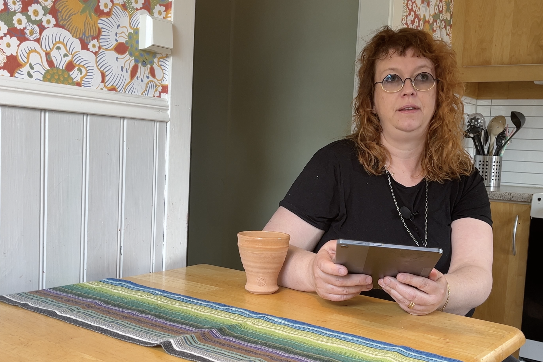 Sjukhusdiakonen Martina Lindberg sitter vid ett köksbord i furu. I handen håller hon en läsplatta med texten till dagens helgmålsbön. Hon har svarta kläder och rött hår. Hennes glasögon är mörka. 