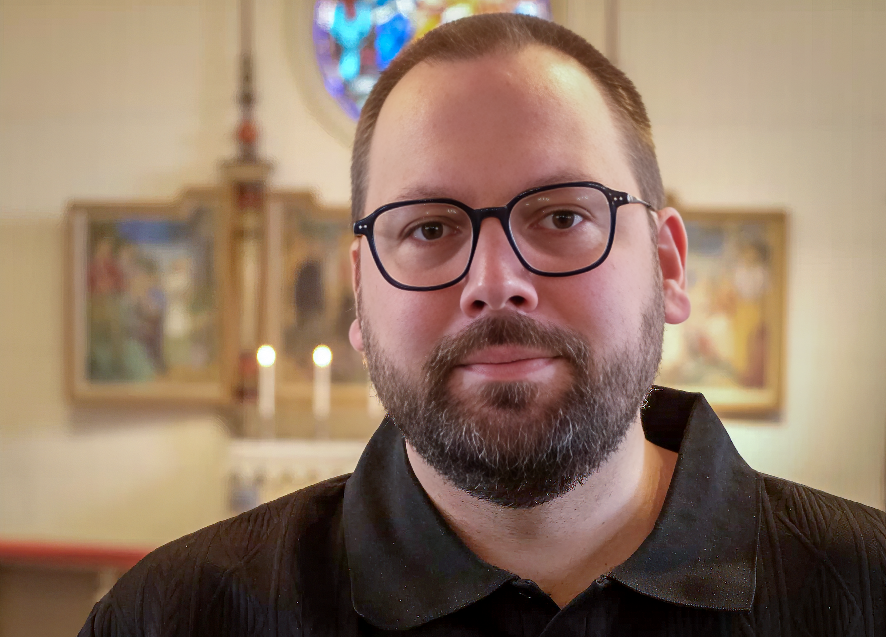 Bilden visar Andreas Forsman ståendes framför altaret i en kyrka. Han har brun tröja och glasögon och helskägg.