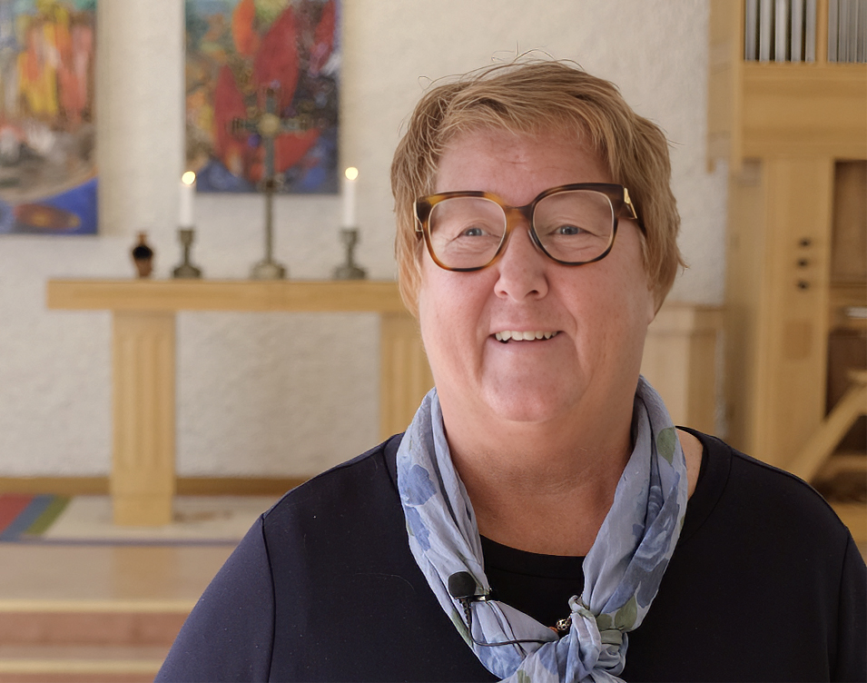 Bilden visar församlingspedagogen Cathrine. Hon står framför altaret i Byskes församlingshem med två ljus och ett krucifix. Cathrine är klädd i en blå tunika med en scarf i en mer ljusblå nyans. Hon har bruna glasögon på sig.