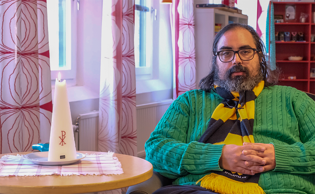 Vi ser församlingspedagogen Erik Belani sittandes i en fåtölj med händerna knäppta. Han bär en grön tröja och en svart/gul AIK-halsduk. 