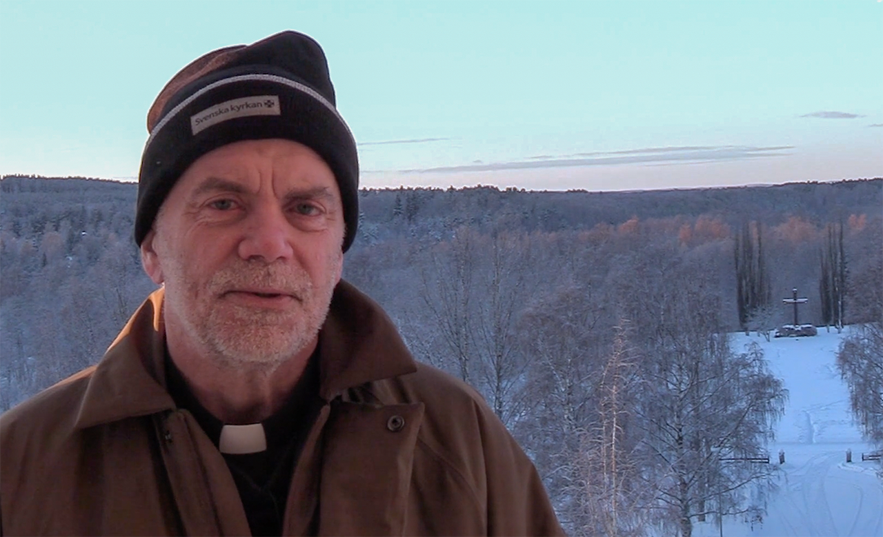 Prästen Arne Andersson i Landskyrkans torn med utsikt mot nordost. Arne håller en digital andakt och är iklädd en brun jacka och en mössa med Svenska kyrkans logotyp 