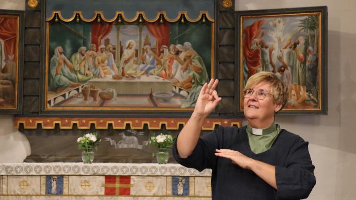 Diakonen Katarina Häggmark använder teckenspråk i Sankt Olovs kyrka i Skellefteå. Katarina står framför altartavlan i kyrkan och är iklädd en svart tunika och grön diakonskjorta. 