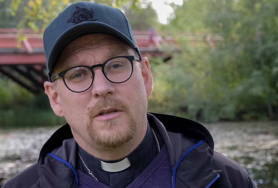Prästen Tom Randgard fotograferad utomhus med keps och vindjacka. Han står framför Lejonströmsbron med sina röda träpiloner.