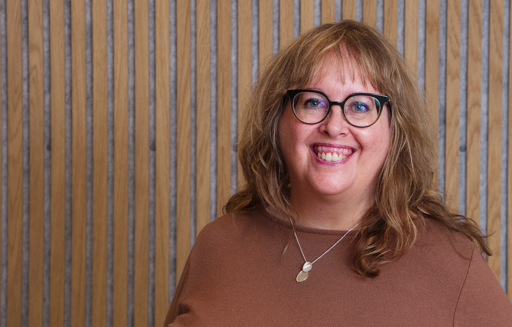 Porträttbild på Ylva Bergström, ambassadör för Hjärnkoll Västerbotten. Hon står framför en ribbad vägg och har brunt långt hår och en brun tröja. Hornbågade glasögon med svart båge.